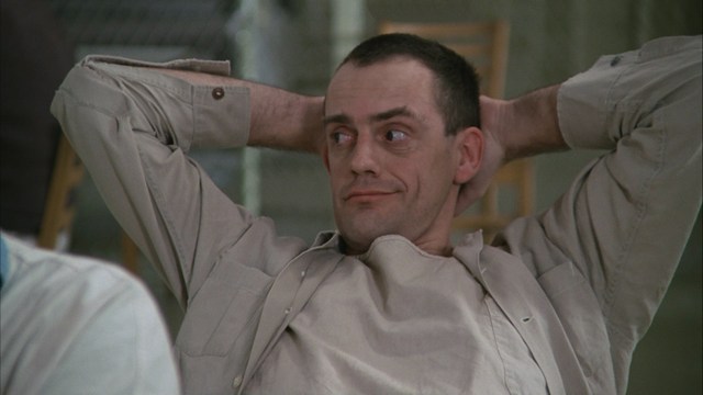 A scene from a movie featuring a man with short black hair and a dark jacket, sitting at a table with a surprised expression, holding a beverage, in a dimly lit indoor setting.