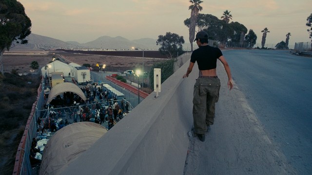 A still from a movie featuring a man standing on a boardwalk in front of an arcade with signs for Skeeball, saltwater taffy, and various attractions.