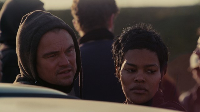 A man with a beard and mustache smiles warmly while standing on a boardwalk by the beach, with colorful flags overhead and a large building in the background.