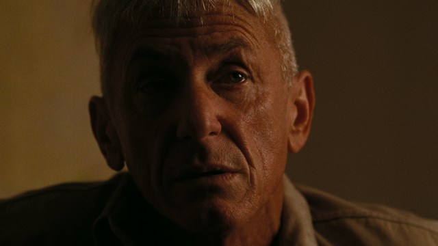 A serious-looking cardinal stands in front of a wooden backdrop with colorful posters related to Catholicism, holding a microphone at a press conference.