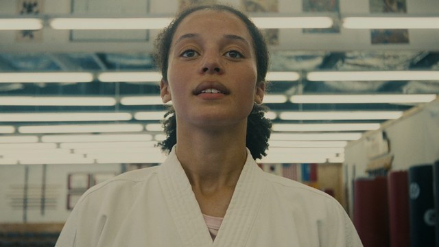 A man in a white suit and hat stands at a doorway, smiling, with a suburban landscape in the background.