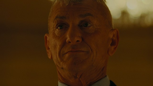 A man with large, feathered angel wings stands in a room, wearing a black suit and looking directly at the viewer.