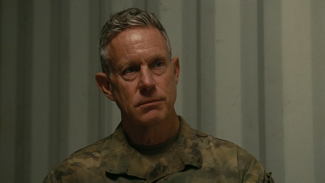 A close-up of a man with a concerned expression, with blurred figures in the background, set in a dimly lit interior.