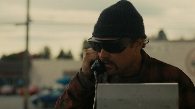 A scene featuring a man with short hair and a beard, wearing a brown jacket and purple shirt, standing outside a restaurant with bright yellow walls and a cartoon cow logo.