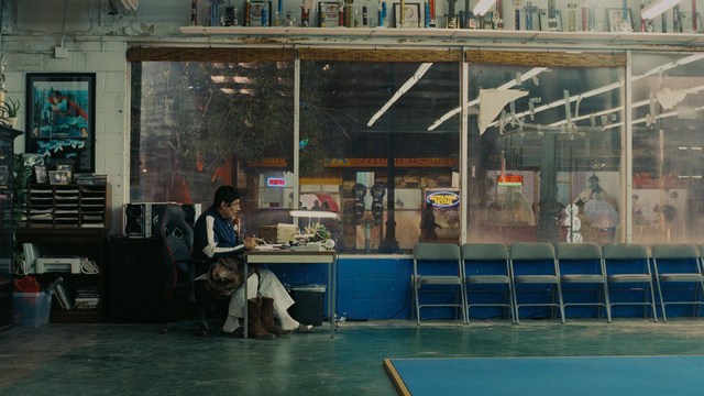 A golden trophy statue of a goblin-like character positioned on a wooden table in an office setting, with large windows and a building facade visible in the background.