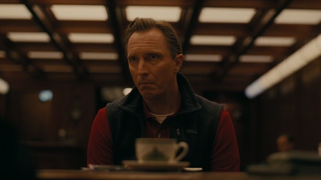 Close-up of a man with a serious expression, seated in a dimly lit area, with soft lighting from background lamps.