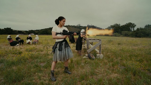 A close-up shot of a young man with short hair and a gray sweatshirt, looking intently at the camera, with a woman sleeping beside him in the background.