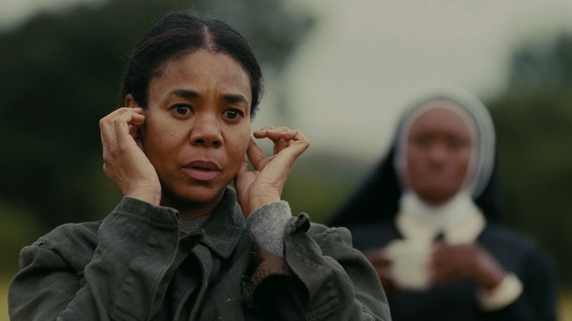 A woman seated at a restaurant table, thoughtfully resting her chin on her hand while reading a newspaper. Blurred figures of two people can be seen in the background.