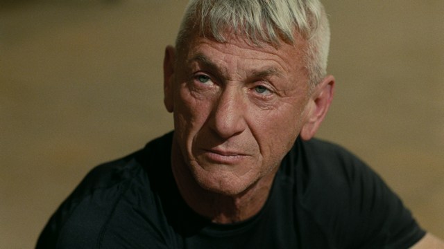 Two characters sitting at a table, one holding a small statue, with a surprised expression. The background features vibrant colors and patterns.