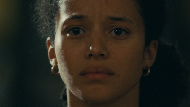 A serious-looking cardinal with a red hat and black robes sitting in a wooden chair in an office, surrounded by colorful posters and memorabilia.