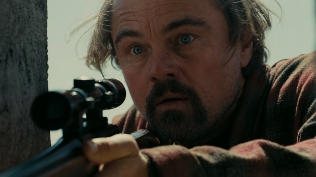 A scene featuring a man in a bishop's attire speaking at a podium surrounded by microphones, with a woman in a red dress beside him and a crowd of onlookers in the background.