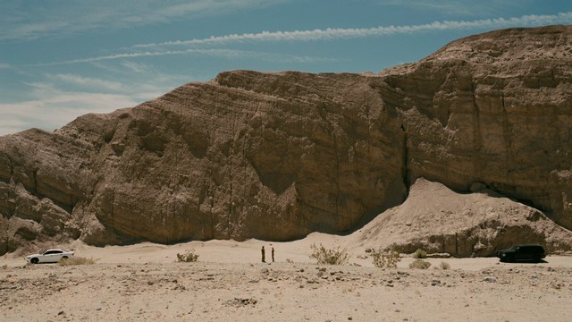 A tense scene featuring a woman with long hair and a serious expression, looking off to the side, while a man with short hair sits in the background, appearing relaxed and slightly turned away.