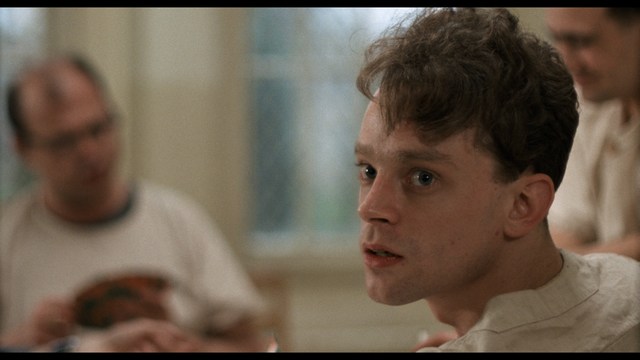 A serious-looking cardinal stands in front of a wooden backdrop with colorful posters related to Catholicism, holding a microphone at a press conference.