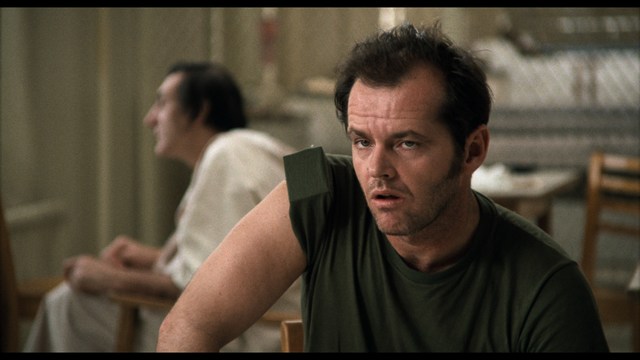 A woman with a pensive expression rests her chin on her hand, sitting at a table with kitchen appliances and items in the background.