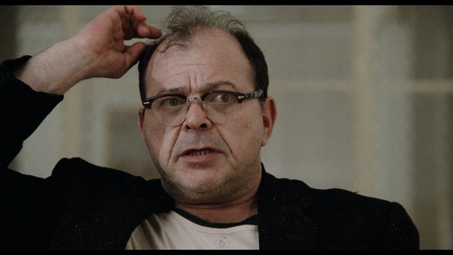 A scene featuring a man with short hair and a beard, wearing a brown jacket and purple shirt, standing outside a restaurant with bright yellow walls and a cartoon cow logo.