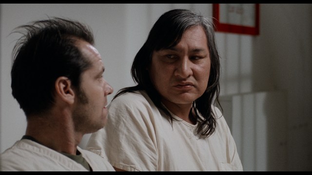 A close-up shot of a man wearing a white suit and a white hat, looking intently at someone in a retail store filled with merchandise.