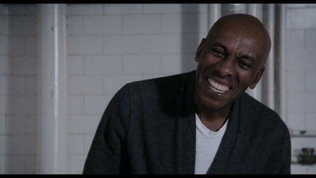 A close-up shot of a young man with short hair and a gray sweatshirt, looking intently at the camera, with a woman sleeping beside him in the background.