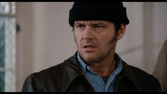 A woman seated at a restaurant table, thoughtfully resting her chin on her hand while reading a newspaper. Blurred figures of two people can be seen in the background.