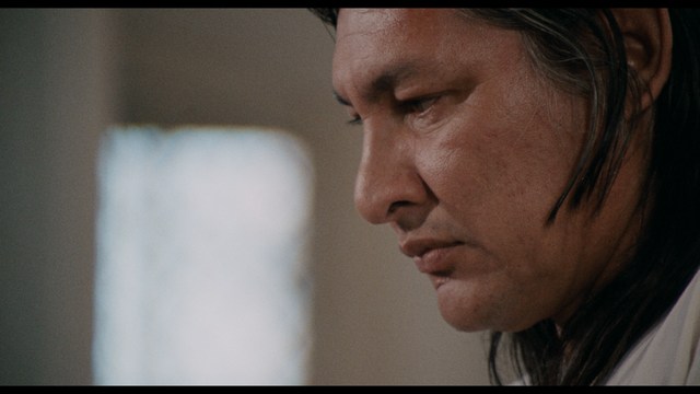 A man with dark, styled hair and a subtle smile seated at a restaurant table, with blurred diners in the background.