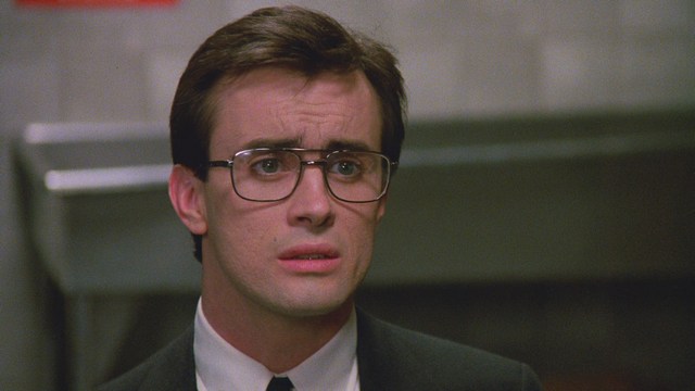 A woman with long dark hair, wearing a brown sweater, gestures with one hand while speaking in a warmly lit room with blinds and colorful artwork in the background.