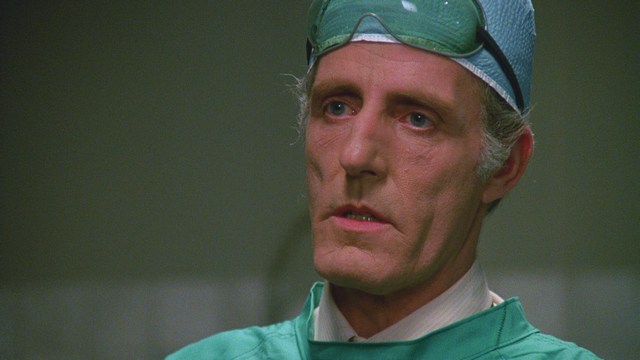 A woman with long dark hair resting her chin on her hand, looking pensively off to the side. In the background, various kitchen appliances and items are visible.