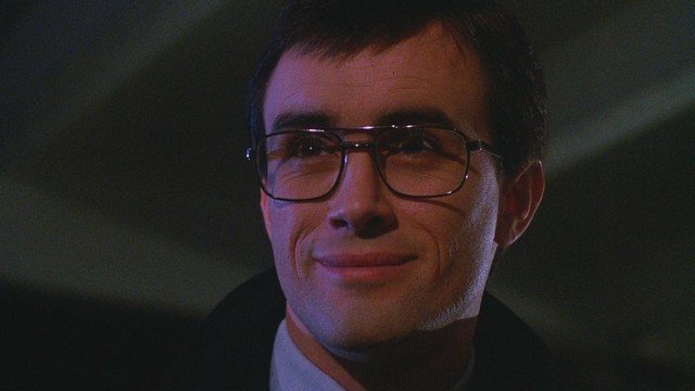 A man in a white suit and hat stands at the entrance of a house, smiling, with a suburban neighborhood visible in the background.