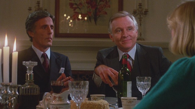 Two men sitting close together in a diner, one wearing a black beanie with the word 'SNOOGANS' and the other wearing a cap, both appearing engaged in conversation.