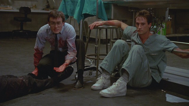 A scene from a movie featuring a man with short black hair and a dark jacket, sitting at a table with a surprised expression, holding a beverage, in a dimly lit indoor setting.