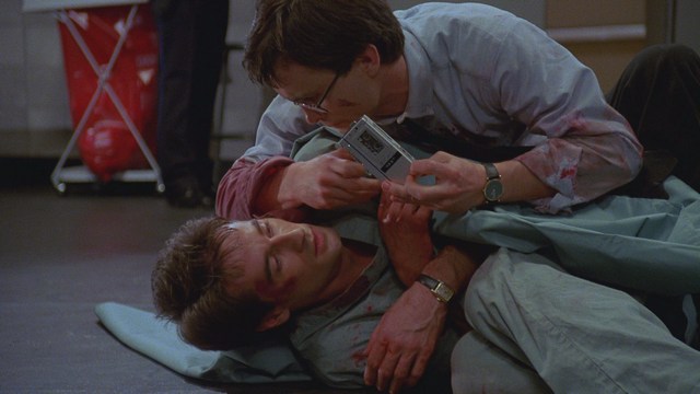 Two male characters in a movie scene, standing indoors near a doorway, with a blurred background of a waiting area.