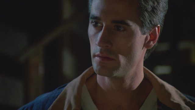 Two male characters in a cozy indoor setting, one with long hair and a beanie, smirking while holding a small object, and the other with a beard and cap, looking surprised. A colorful background with posters can be seen.