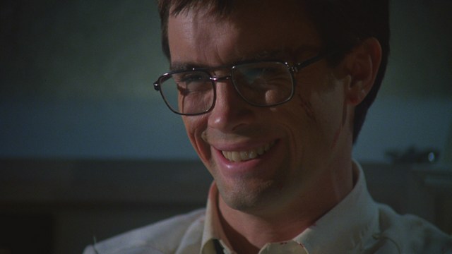 A close-up of a man wearing a red cardinal's hat and black clerical robes, seated in a leather armchair with a serious expression, in an office setting adorned with colorful posters.