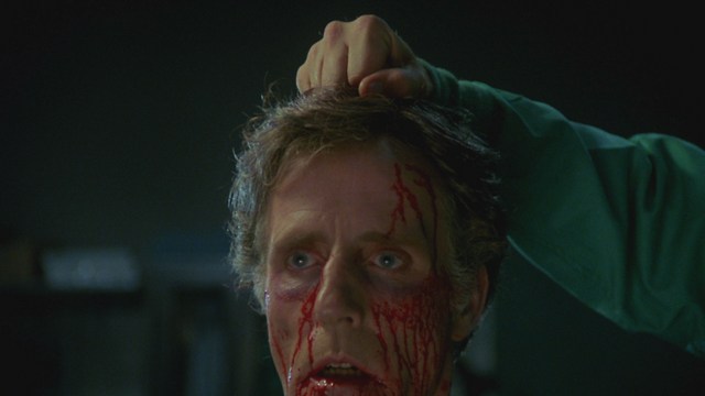 A scene from a movie featuring a man dressed in a bishop's robe speaking at a podium surrounded by microphones, with a woman in a red outfit smiling next to him, and an audience in the background.