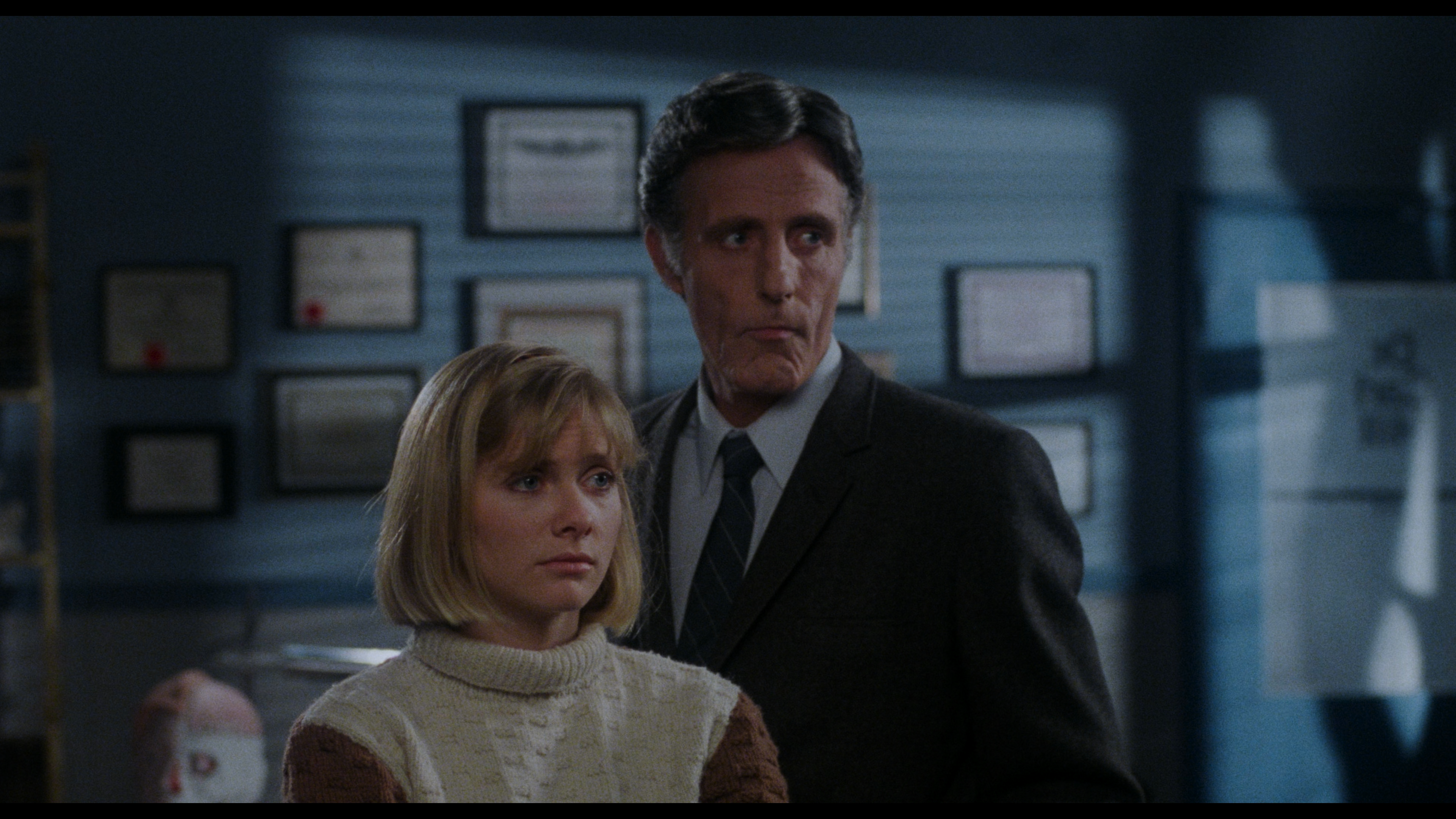 A man wearing a white hat and a light-colored suit looks intently at someone, standing inside a store filled with merchandise and posters.