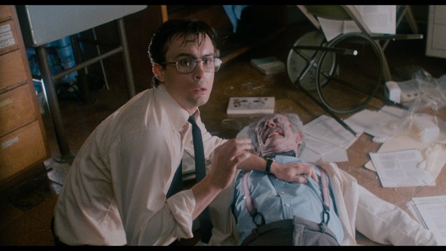 A man with a beard and mustache smiles warmly while standing on a boardwalk by the beach, with colorful flags overhead and a large building in the background.