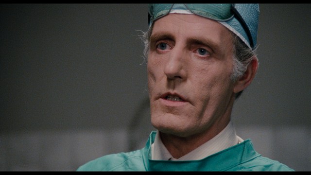 A woman with a pensive expression rests her chin on her hand, sitting at a table with kitchen appliances and items in the background.