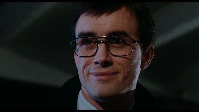 A man in a white suit and hat stands at a doorway, smiling, with a suburban landscape in the background.