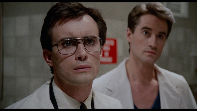 A man with large, feathered angel wings stands in a room, wearing a black suit and looking directly at the viewer.