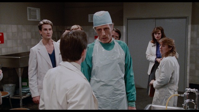 A close-up of a man with a concerned expression, with blurred figures in the background, set in a dimly lit interior.