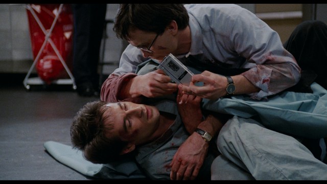 Two young men are looking at something off-camera, standing inside a building with bright lighting and seating areas visible in the background.