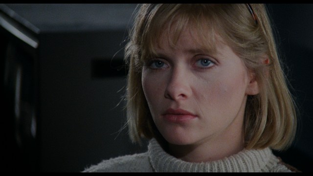 Close-up of a man with a serious expression, seated in a dimly lit area, with soft lighting from background lamps.