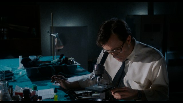 A close-up shot of a young man with short hair and a gray sweatshirt, looking intently at the camera, with a woman sleeping beside him in the background.