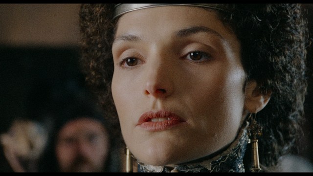 A tense scene featuring a woman with long hair and a serious expression, looking off to the side, while a man with short hair sits in the background, appearing relaxed and slightly turned away.