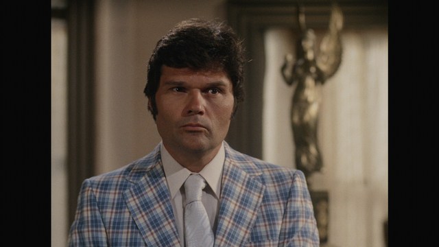 A man wearing a white suit and hat appears to be having a serious conversation in a retail store, with various products displayed in the background.