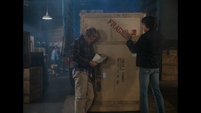 Two men examining a large wooden crate marked 'FRAGILE' in a warehouse setting, surrounded by wooden boxes and dim lighting.