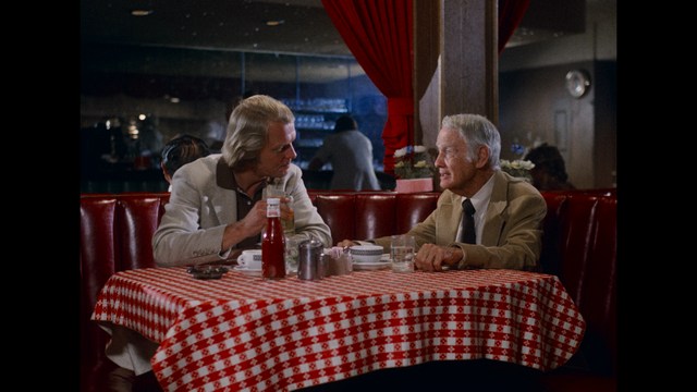 Two men sitting at a red vinyl booth in a diner, conversing over a checkered tablecloth with condiments visible.