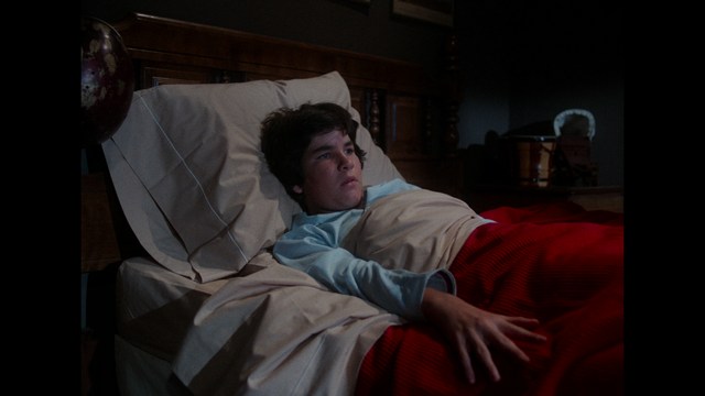 A young person lying in bed, looking pensive and awake, with light-colored bedding and a dark room background.