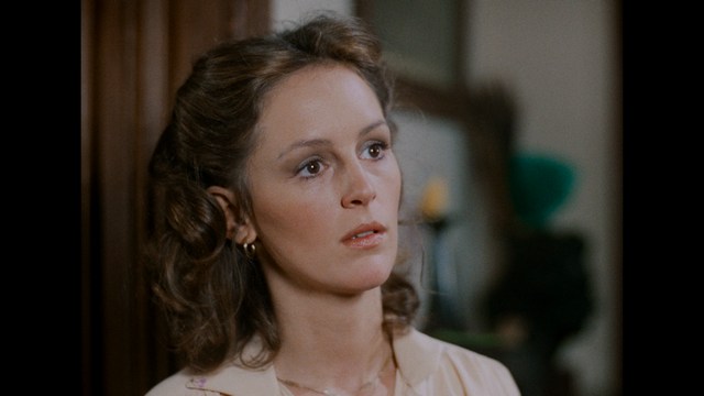 A woman with shoulder-length curly hair looks thoughtfully, wearing a light-colored shirt in an indoor setting.