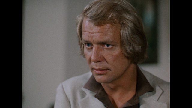 Close-up of a man with light brown hair, wearing a light-colored blazer and a dark shirt, looking intently with an expression of concern or contemplation.