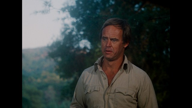 A police officer examines a small object while a man in a green jacket looks on, both standing in a grassy area with trees in the background.