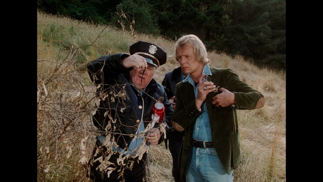 Two men, one in a police uniform and the other in casual attire, examining a plant in an outdoor grassy area.